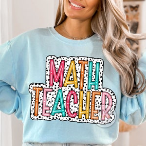 Op de afbeelding: Een lichtblauw sweatshirt met de tekst "MATH TEACHER" in een kleurrijk, omlijnd lettertype met zwarte stippen.