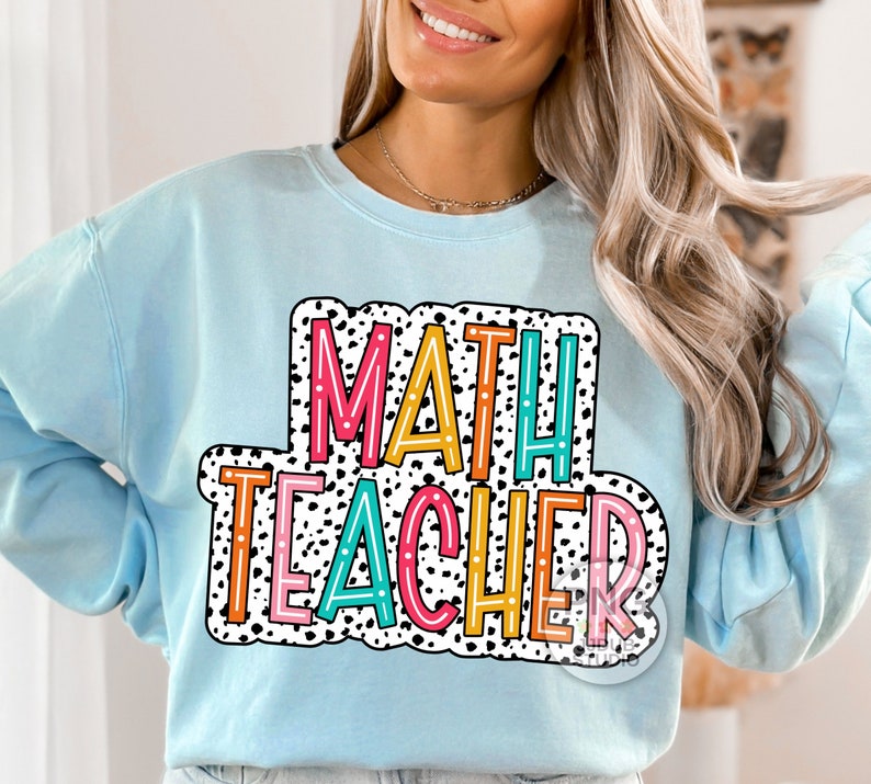 Op de afbeelding: Een lichtblauw sweatshirt met de tekst "MATH TEACHER" in een kleurrijk, omlijnd lettertype met zwarte stippen.
