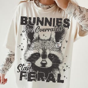 K&ouml;nnte beinhalten: Ein wei&szlig;es T-Shirt mit einer schwarzen Grafik eines Waschb&auml;ren mit dem Text "Bunnies Are Overrated Stay Feral".