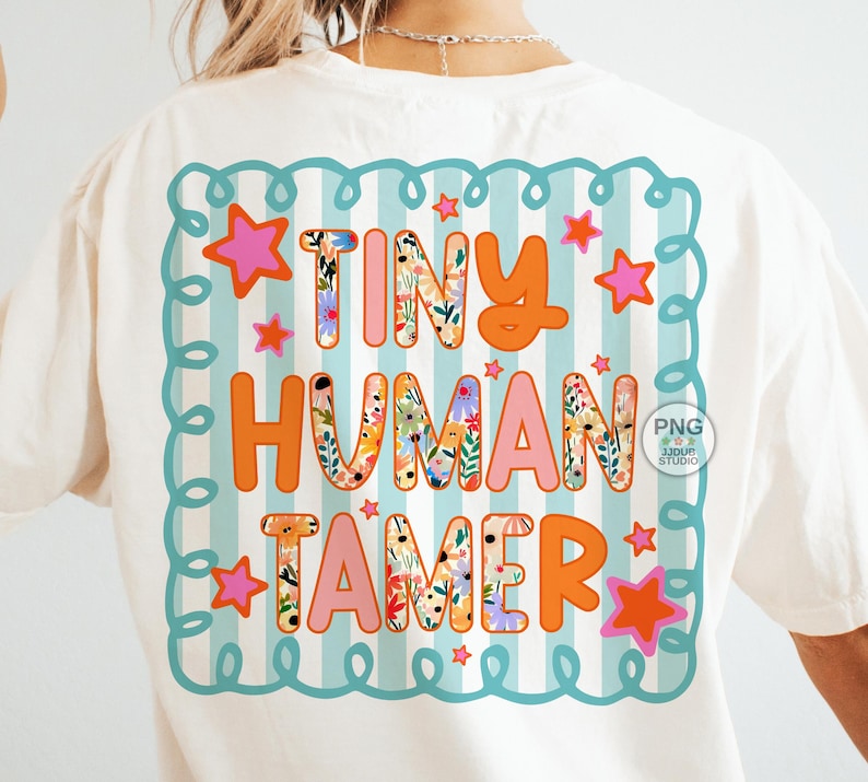 以下が含まれることがあります： クリーム色のTシャツにカラフルなグラフィック。グラフィックは、オレンジとピンクで「TINY HUMAN TAMER」という言葉を特徴とし、文字には花のパターンがあります。デザインは星が付いたターコイズのボーダーで囲まれています。