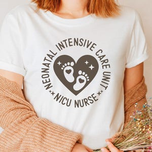 May include: White t-shirt featuring a circular design with the text "Neonatal Intensive Care Unit NICU Nurse." The design includes a heart shape with baby footprints and stars. The shirt is a casual style.