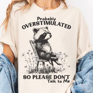 K&ouml;nnte beinhalten: Ein beiges T-Shirt mit einer schwarz-wei&szlig;en Illustration eines Waschb&auml;ren, der auf einem Stuhl sitzt. Der Text "Probably Overstimulated So Please Don't Talk to Me" ist auf dem T-Shirt aufgedruckt.