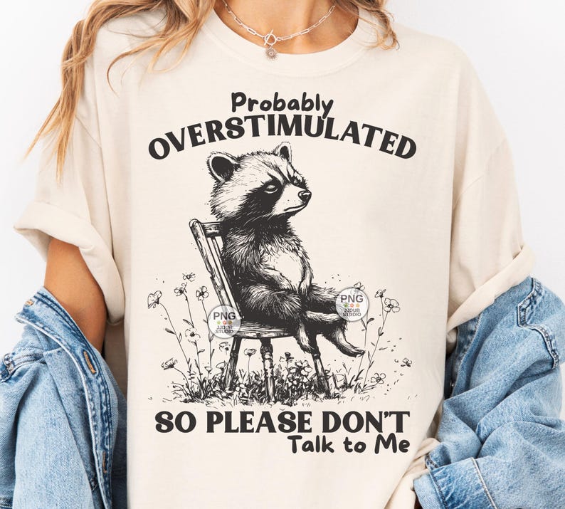 K&ouml;nnte beinhalten: Ein beiges T-Shirt mit einer schwarz-wei&szlig;en Illustration eines Waschb&auml;ren, der auf einem Stuhl sitzt. Der Text "Probably Overstimulated So Please Don't Talk to Me" ist auf dem T-Shirt aufgedruckt.