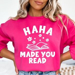 May include: A pink sweatshirt with the text "HAHA MADE YOU READ" in white. The text is surrounded by a stylized open book and small white flowers.