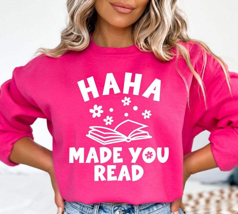 May include: A pink sweatshirt with the text "HAHA MADE YOU READ" in white. The text is surrounded by a stylized open book and small white flowers.