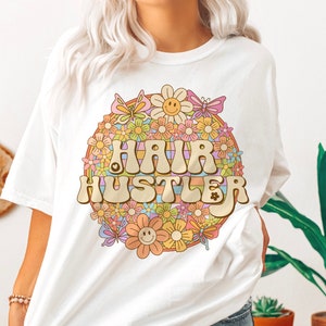 May include: White t-shirt with a retro-style graphic of a rainbow circle with flowers and butterflies surrounding the text "HAIR HUSTLER".