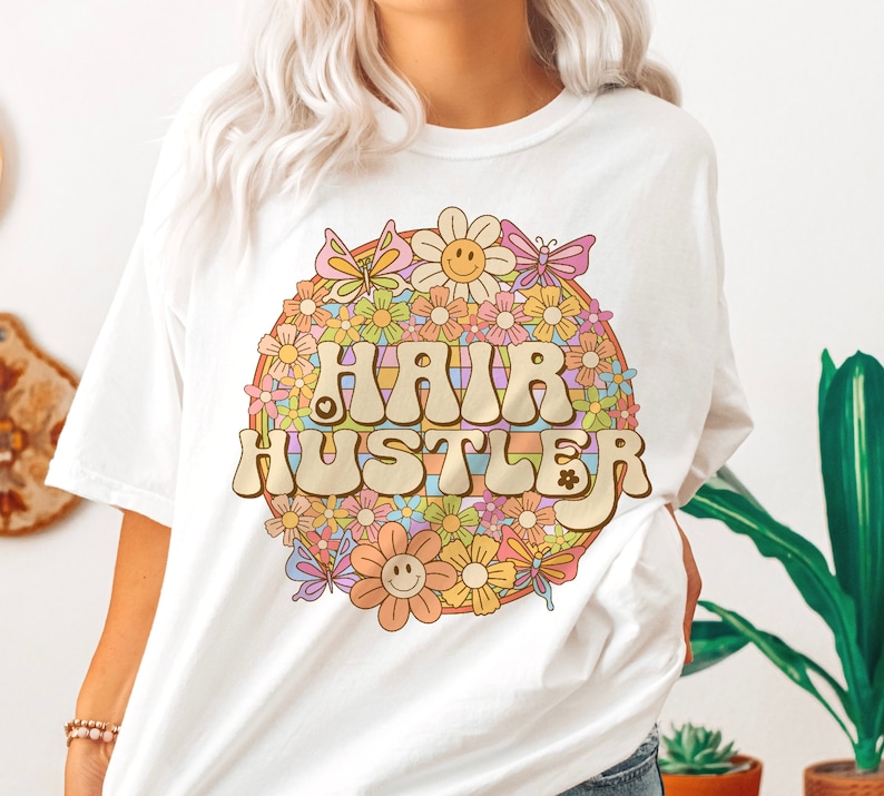 May include: White t-shirt with a retro-style graphic of a rainbow circle with flowers and butterflies surrounding the text "HAIR HUSTLER".