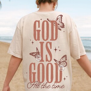 May include: A cream-coloured t-shirt featuring the text "GOD IS GOOD All the time" in a rust-coloured font. The design incorporates butterfly illustrations and star accents, giving a vintage feel. The t-shirt has a relaxed fit.