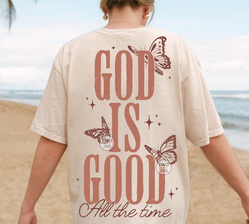 May include: A cream-coloured t-shirt featuring the text "GOD IS GOOD All the time" in a rust-coloured font. The design incorporates butterfly illustrations and star accents, giving a vintage feel. The t-shirt has a relaxed fit.