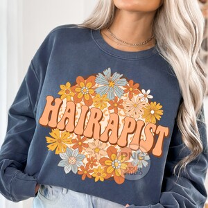 May include: A dark blue sweatshirt with a retro-style graphic of the word "HAIRAPIST" surrounded by colorful flowers. The flowers are in shades of orange, yellow, blue, and green.