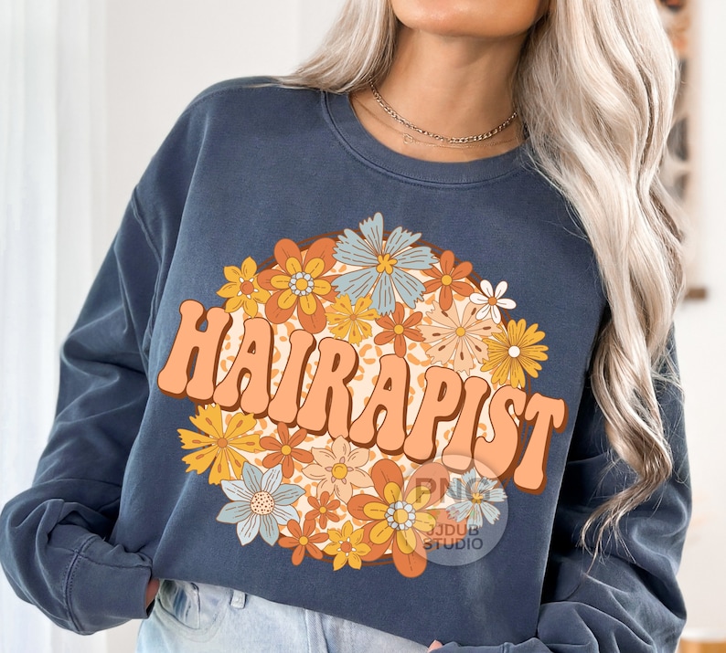 May include: A dark blue sweatshirt with a retro-style graphic of the word "HAIRAPIST" surrounded by colorful flowers. The flowers are in shades of orange, yellow, blue, and green.
