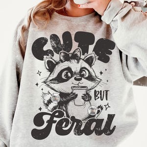 K&ouml;nnte beinhalten: Ein graues Sweatshirt mit einer schwarz-wei&szlig;en Illustration eines Waschb&auml;ren, der eine Tasse h&auml;lt. Der Text "Cute but Feral" ist auf dem Sweatshirt aufgedruckt.