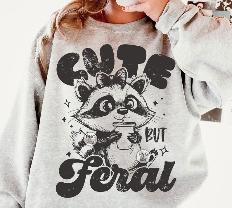 K&ouml;nnte beinhalten: Ein graues Sweatshirt mit einer schwarz-wei&szlig;en Illustration eines Waschb&auml;ren, der eine Tasse h&auml;lt. Der Text "Cute but Feral" ist auf dem Sweatshirt aufgedruckt.