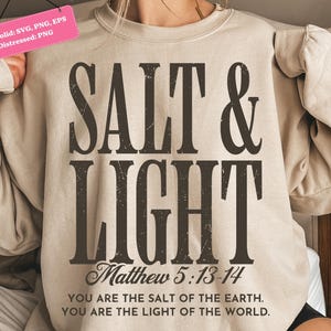 Könnte beinhalten: Beiges Sweatshirt mit dem Text "SALT & LIGHT" in großen, verwitterten, dunkelbraunen Buchstaben. Darunter steht "Matthew 5:13-14" und ein Zitat. Das lässige Kleidungsstück soll inspirieren und erheben.