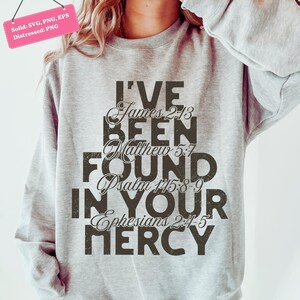 以下が含まれることがあります： 「I'VE BEEN FOUND IN YOUR MERCY」の文字が、くすんだ濃い茶色のフォントでプリントされたライトグレーのスウェットシャツ。聖書の詩篇、ヤコブ2:13、マタイ5:7、詩篇145:8-9、エペソ2:4-5が含まれています。左上隅にあるピンク色のタグには「Solid: SVG, PNG, EPS Distressed: PNG」と書かれています。