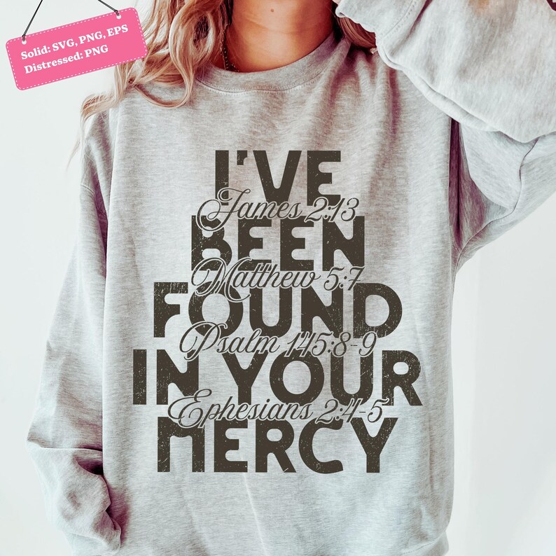 以下が含まれることがあります： 「I'VE BEEN FOUND IN YOUR MERCY」の文字が、くすんだ濃い茶色のフォントでプリントされたライトグレーのスウェットシャツ。聖書の詩篇、ヤコブ2:13、マタイ5:7、詩篇145:8-9、エペソ2:4-5が含まれています。左上隅にあるピンク色のタグには「Solid: SVG, PNG, EPS Distressed: PNG」と書かれています。