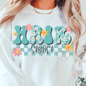 May include: A white sweatshirt with a blue and white floral design that says "Hair Stylist" in a retro font. The design is on a checkered background with pink and yellow flowers.