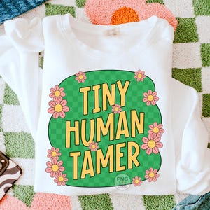 Peut inclure: Sweat-shirt blanc avec un motif circulaire vert et &agrave; carreaux. Le cercle contient le texte "Tiny Human Tamer" en lettres jaunes et des fleurs roses autour du bord.