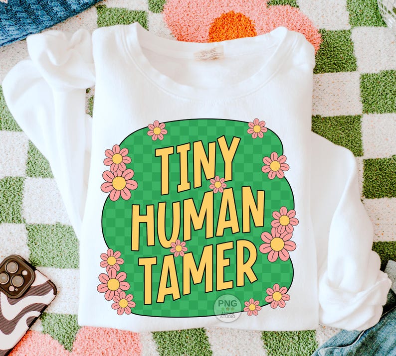Peut inclure: Sweat-shirt blanc avec un motif circulaire vert et &agrave; carreaux. Le cercle contient le texte "Tiny Human Tamer" en lettres jaunes et des fleurs roses autour du bord.