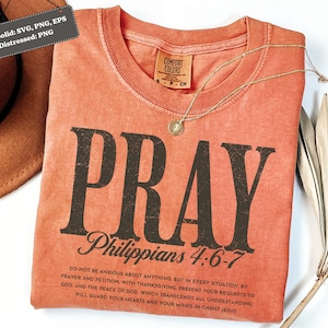 Op de afbeelding: Een koraalkleurig t-shirt met het woord "PRAY" in grote, verweerde zwarte letters. Daaronder staat de tekst "Filippenzen 4:6-7" en een langere passage. Een gouden ketting met een hanger ligt over het shirt. Het shirt is van het merk Comfort Colors.