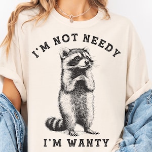 May include: A beige t-shirt featuring a black and white illustration of a raccoon. The text "I'm not needy I'm wanty" is printed on the shirt.