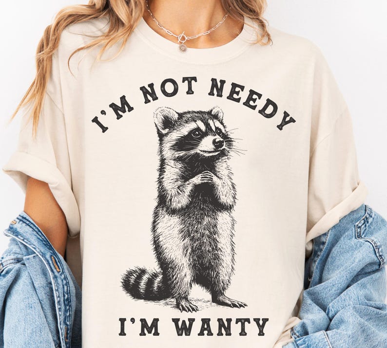 May include: A beige t-shirt featuring a black and white illustration of a raccoon. The text "I'm not needy I'm wanty" is printed on the shirt.
