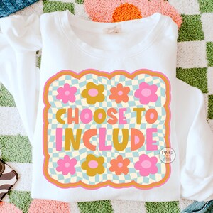 May include: White sweatshirt with a retro design that says "CHOOSE TO INCLUDE". The design features a checkered background, flowers, and a wavy border in shades of pink, orange, and yellow. The sweatshirt is on a colorful patterned surface.