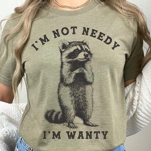 May include: A green t-shirt featuring a raccoon graphic and the text "I'm not needy I'm wanty".
