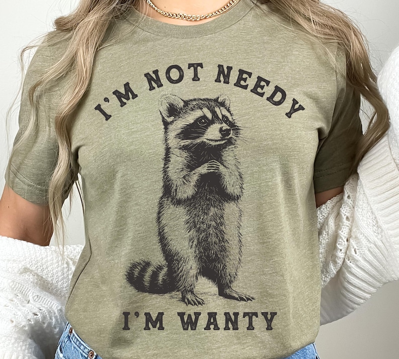 May include: A green t-shirt featuring a raccoon graphic and the text "I'm not needy I'm wanty".
