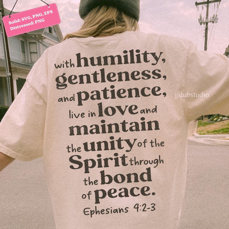以下が含まれることがあります： 黒いテキストデザインのクリーム色のTシャツ。テキストには「謙虚さ、優しさ、忍耐、愛、統一、精神、絆、平和」という言葉と聖書の節エペソ4：2-3が含まれています。シャツは人が着ています。