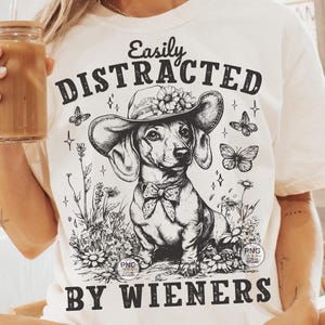 May include: White t-shirt with a black and white illustration of a dachshund wearing a flower crown and the text "Easily Distracted By Wieners".