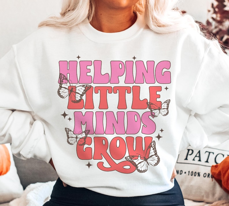 Peut inclure: Sweat-shirt blanc avec un texte rose et rouge qui dit "Helping Little Minds Grow" avec des motifs de papillons.