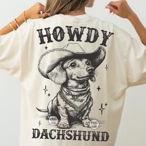 May include: A cream-colored t-shirt with a black and white illustration of a dachshund wearing a cowboy hat. The text "HOWDY DACHSHUND" is printed on the shirt.