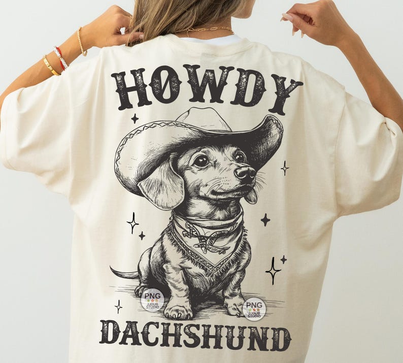 May include: A cream-colored t-shirt with a black and white illustration of a dachshund wearing a cowboy hat. The text "HOWDY DACHSHUND" is printed on the shirt.