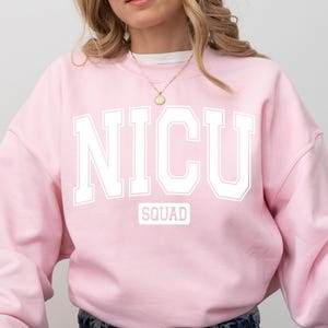 May include: A light pink sweatshirt with the text "NICU SQUAD" in white block letters. The word "SQUAD" is in a smaller white rectangle below the main text. The sweatshirt has long sleeves and a crew neck.