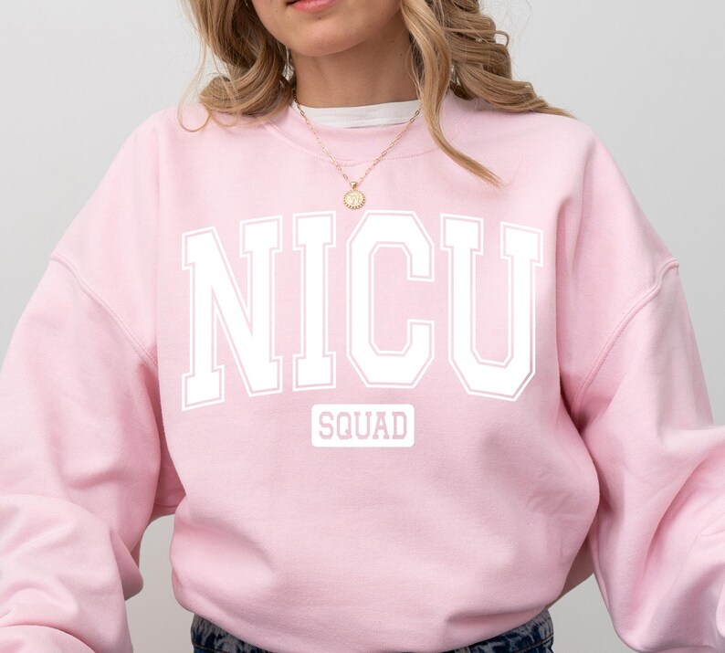 May include: A light pink sweatshirt with the text "NICU SQUAD" in white block letters. The word "SQUAD" is in a smaller white rectangle below the main text. The sweatshirt has long sleeves and a crew neck.