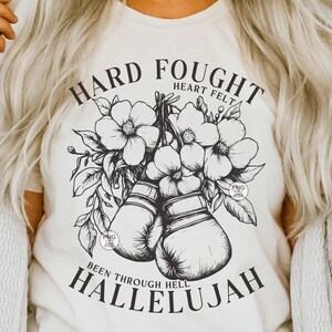 Peut inclure: Un t-shirt blanc avec un motif graphique noir repr&eacute;sentant des gants de boxe, des fleurs et le texte "Hard Fought Heart Felt Been Through Hell Hallelujah".
