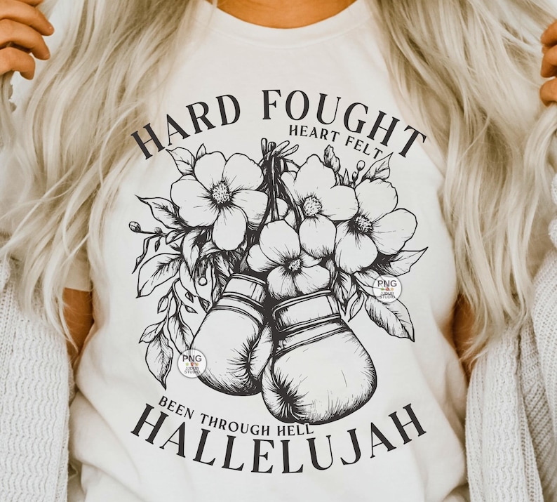 Peut inclure: Un t-shirt blanc avec un motif graphique noir repr&eacute;sentant des gants de boxe, des fleurs et le texte "Hard Fought Heart Felt Been Through Hell Hallelujah".