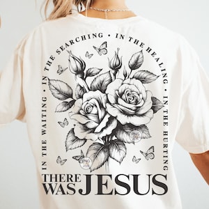 Peut inclure: Un t-shirt blanc avec un motif graphique noir repr&eacute;sentant un bouquet de roses et de papillons. Le texte "IN THE WAITING. IN THE SEARCHING. IN THE HEALING. IN THE HURTING. THERE WAS JESUS" entoure les roses.