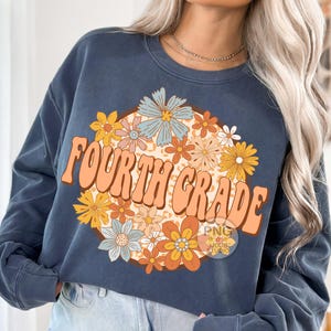 May include: A navy blue sweatshirt with a graphic design of a circle of colorful flowers and the text "FOURTH GRADE" in a retro font.