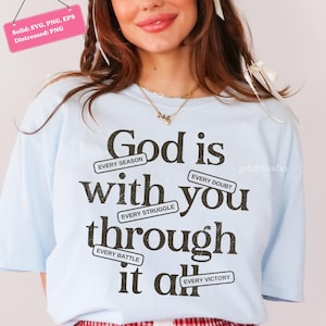 May include: Light blue t-shirt with the message "God is with you through it all" in black text. The text includes phrases like "Every Season," "Every Doubt," and "Every Victory." The shirt has a distressed design.