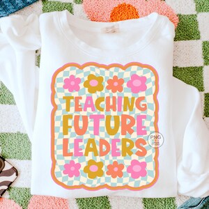 以下が含まれることがあります： オレンジ、ピンク、黄色の「TEACHING FUTURE LEADERS」の文字が特徴的な白いスウェットシャツ。花柄とチェック柄のデザインが施されています。スウェットシャツは模様のある表面に折りたたまれています。レトロな雰囲気のデザインです。