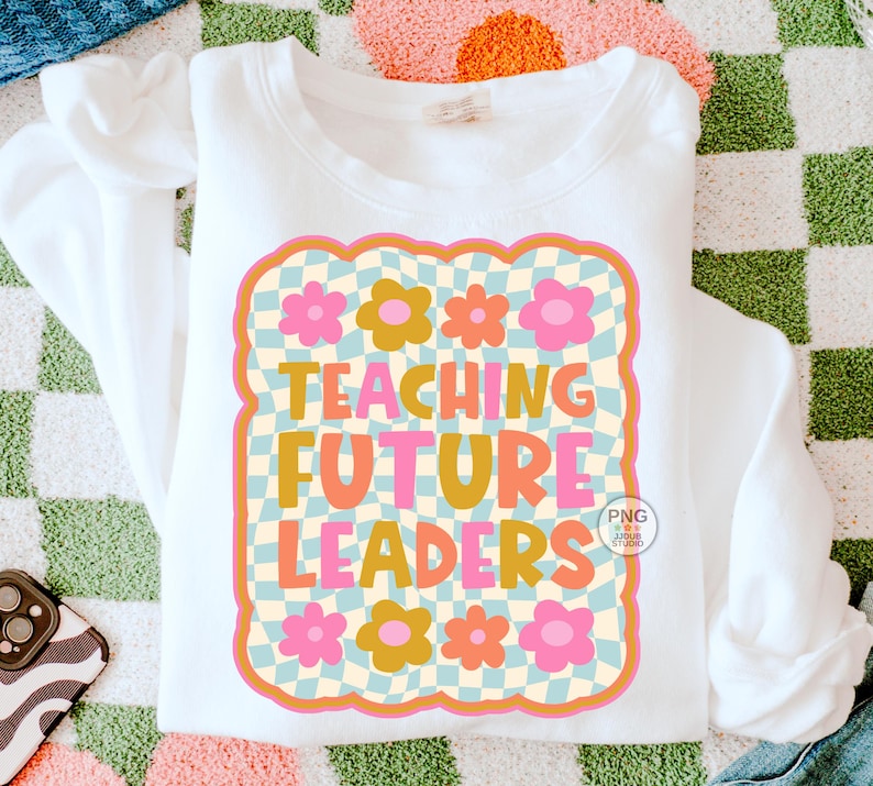 以下が含まれることがあります： オレンジ、ピンク、黄色の「TEACHING FUTURE LEADERS」の文字が特徴的な白いスウェットシャツ。花柄とチェック柄のデザインが施されています。スウェットシャツは模様のある表面に折りたたまれています。レトロな雰囲気のデザインです。