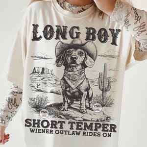 May include: A white t-shirt with a black and white illustration of a dachshund wearing a cowboy hat and bandana. The text "Long Boy Short Temper Wiener Outlaw Rides On" is printed on the shirt.