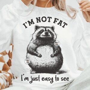 K&ouml;nnte beinhalten: Wei&szlig;es T-Shirt mit einer schwarz-wei&szlig;en Illustration eines Waschb&auml;ren. Der Text "Ich bin nicht dick, ich bin einfach leicht zu sehen" ist auf das T-Shirt gedruckt.