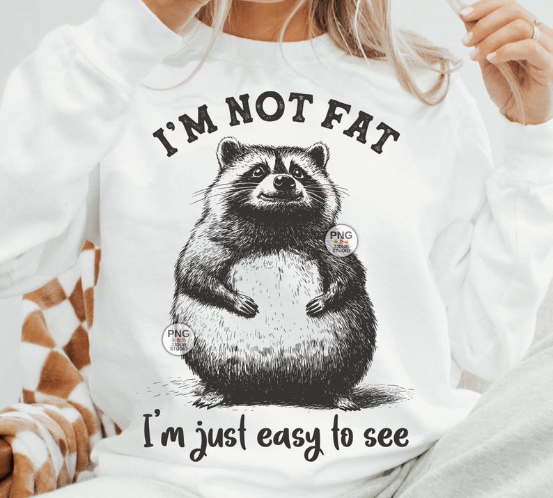 K&ouml;nnte beinhalten: Wei&szlig;es T-Shirt mit einer schwarz-wei&szlig;en Illustration eines Waschb&auml;ren. Der Text "Ich bin nicht dick, ich bin einfach leicht zu sehen" ist auf das T-Shirt gedruckt.