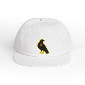 Puede incluir: Una gorra de béisbol blanca con un parche bordado de un pájaro negro de perfil. El pájaro tiene un contorno amarillo y un pequeño corazón rojo con una corona dorada en el pecho. La gorra presenta una corona estructurada y visera plana.