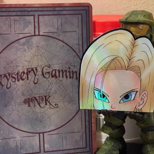 May include: A holographic sticker featuring a cartoon anime character with blonde hair and blue eyes. The character is looking to the left. The sticker is on a green background.