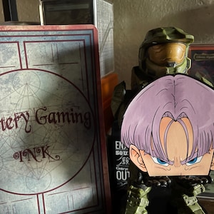 May include: A purple-haired anime character cutout with blue eyes is in front of a green military figure. A sign with the text "Mystery Gaming Ink" is on the left. A card is visible on the right.