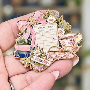 May include: A gold-toned enamel pin featuring a library card with the text LIBRARY CARD, DATE DUE, and BORROWER. The design includes a pink bow, stacked books in green, pink, and blue, white daisies, a floral teacup, and round wire-rimmed glasses.